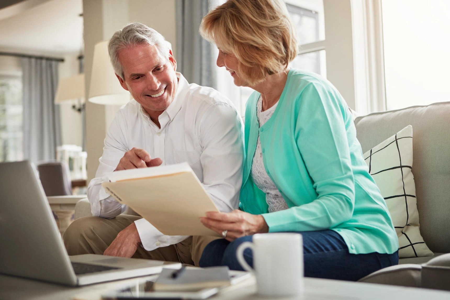 Older couple reviewing paperwork together on sofa with laptop nearby.