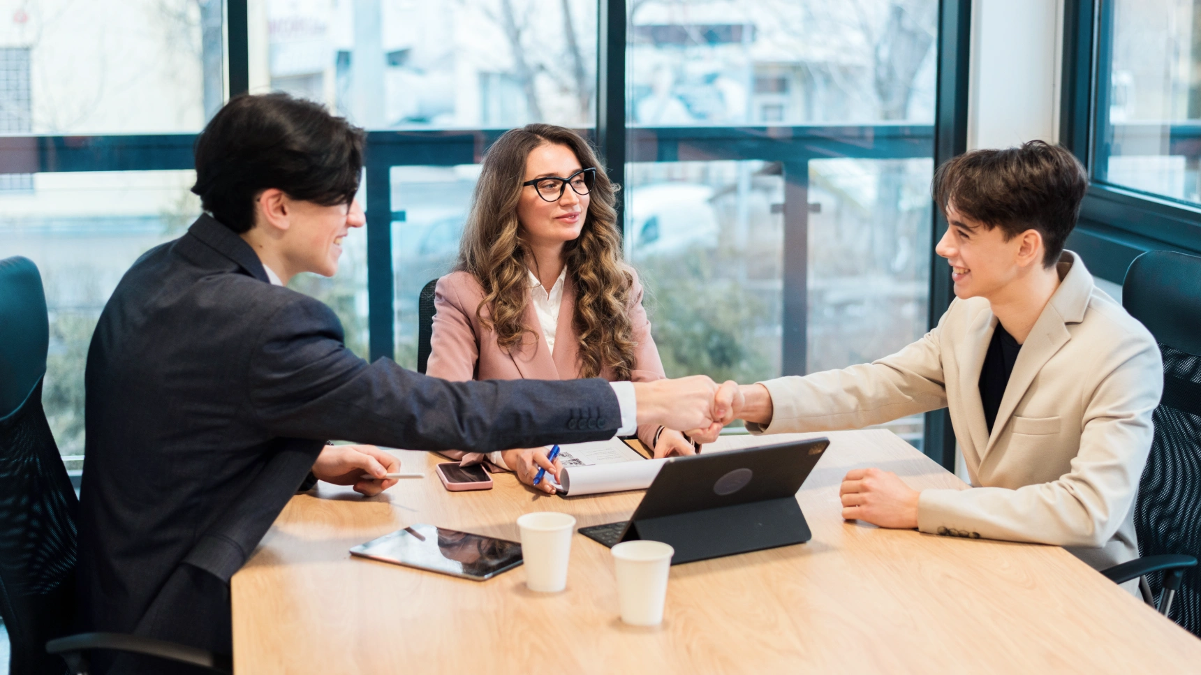 Two young businessmen shaking hands while colleague observes across conference table.