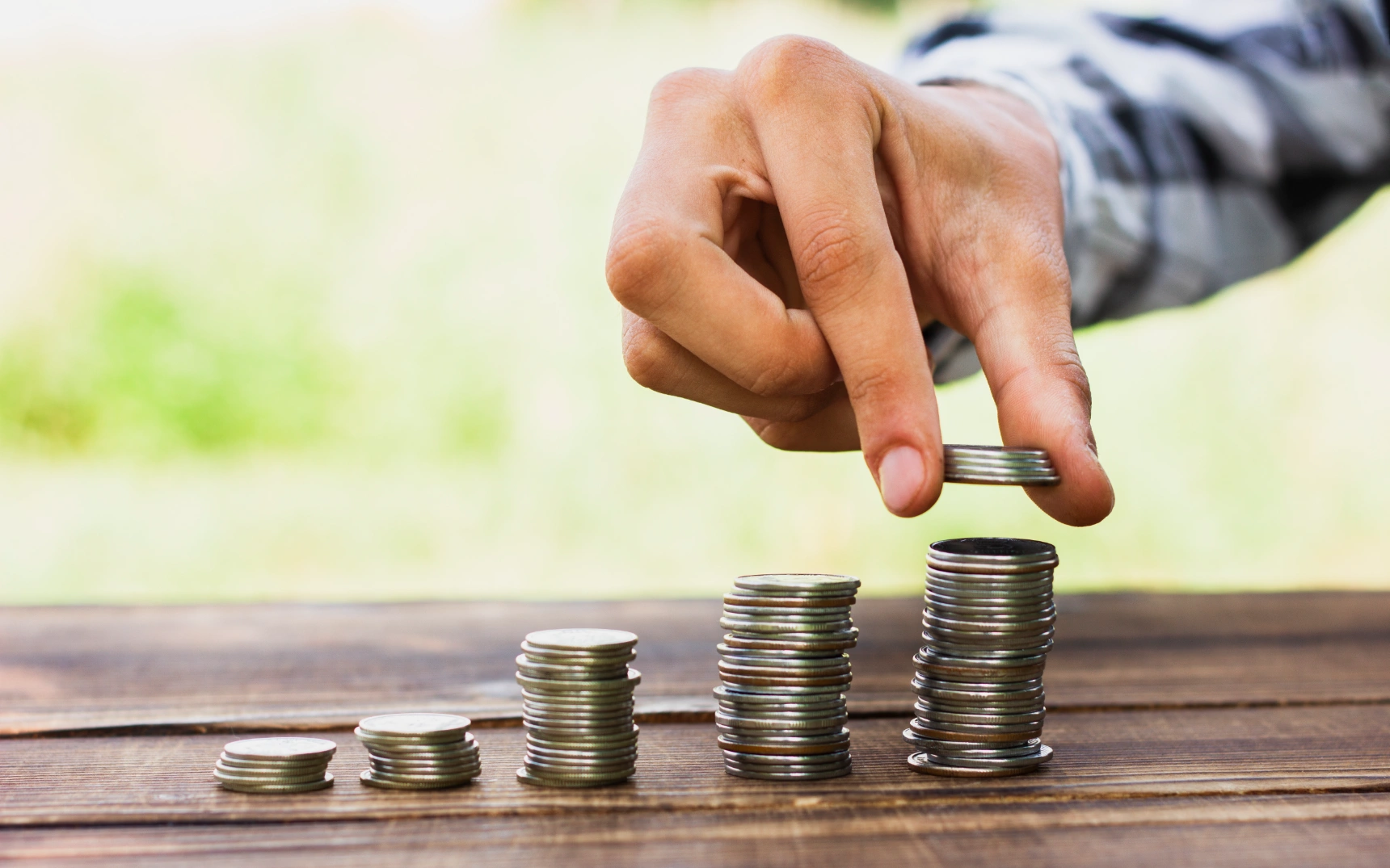 Hand stacking ascending coin piles on wooden table outdoors, representing savings.