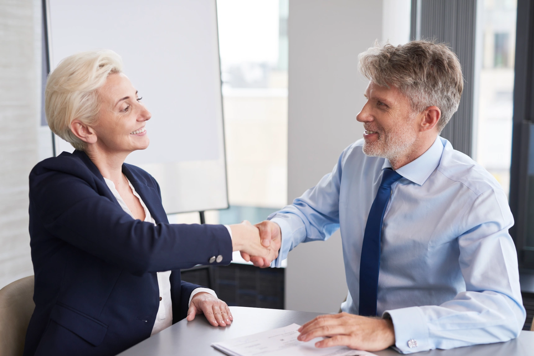 Mature businesswoman and businessman warmly shaking hands during office meeting.